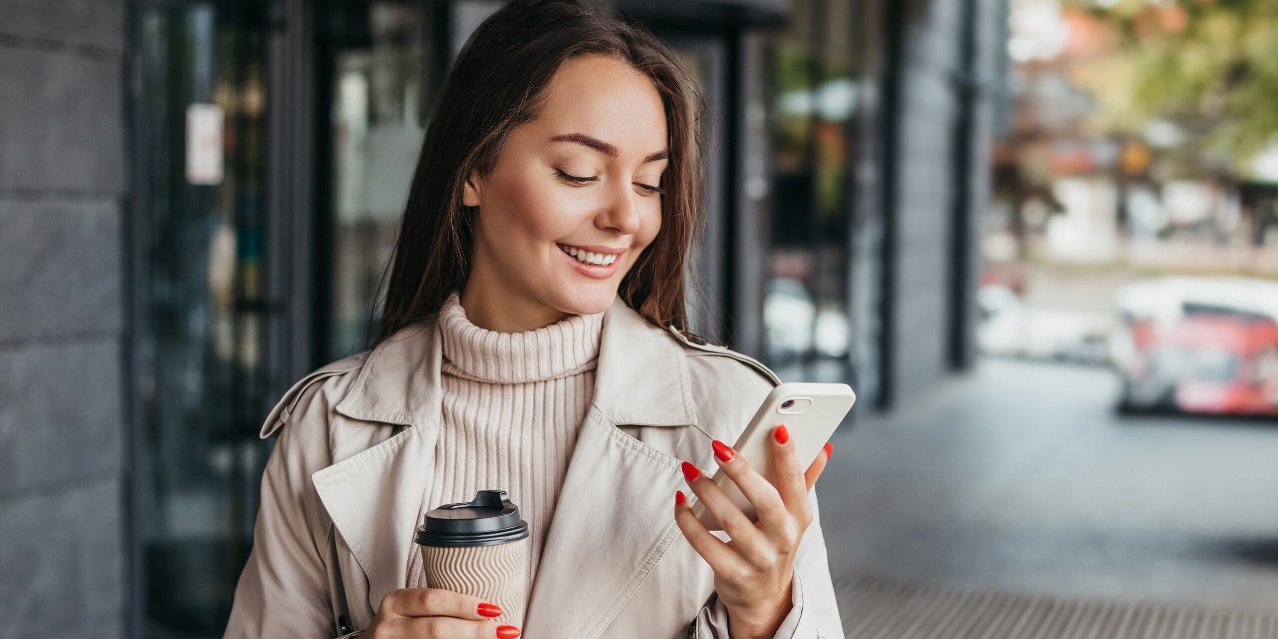 Young smiling ukrainian brunette woman holding a smartphone and a cup of coffee looking at device screen against the background of an office building
