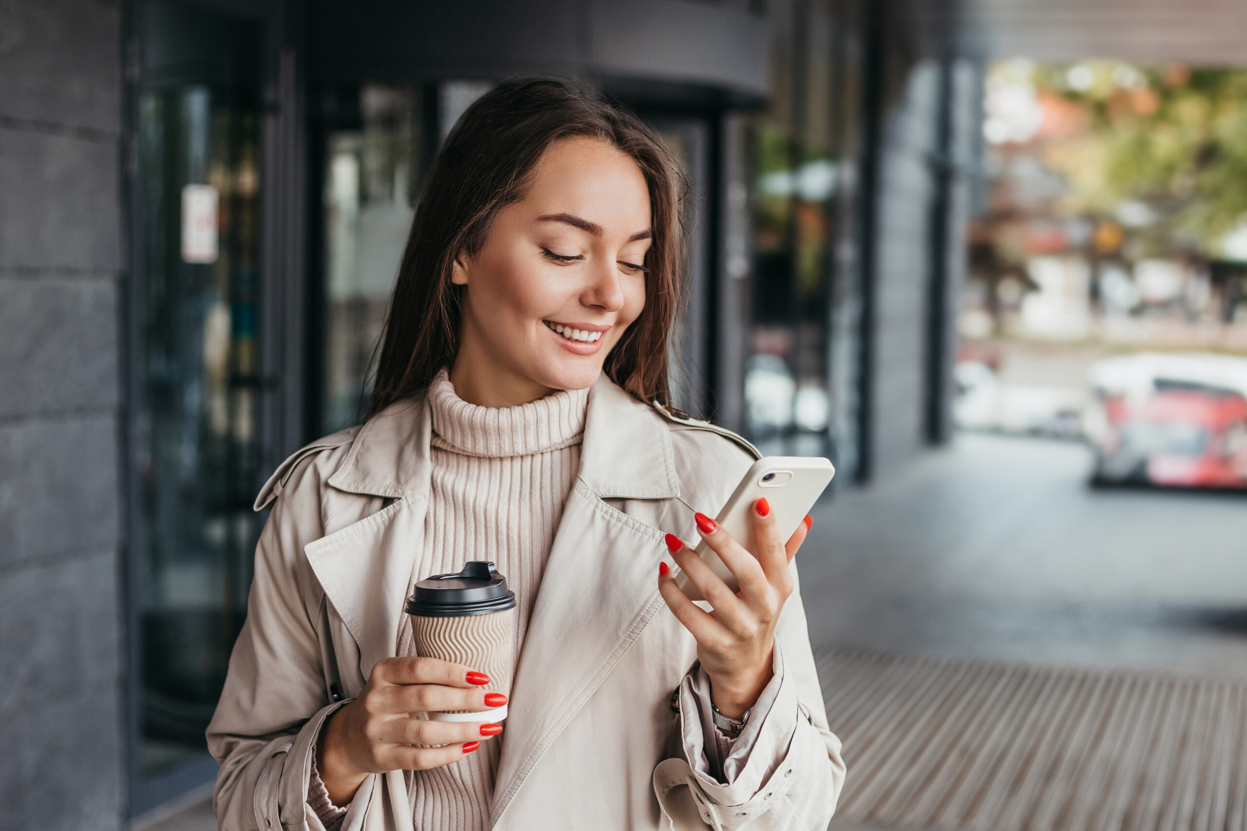 Young smiling ukrainian brunette woman holding a smartphone and a cup of coffee looking at device screen against the background of an office building